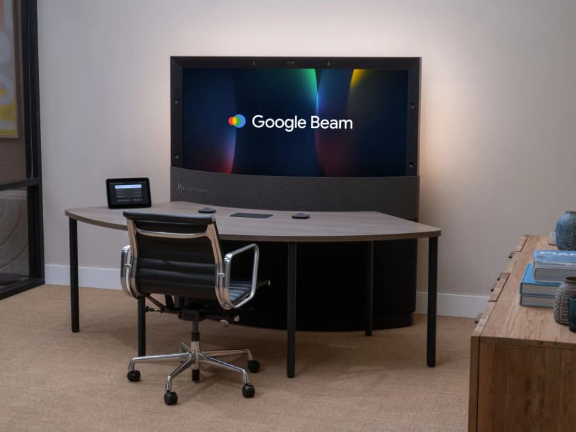 白い壁の小さな部屋に置かれたHP Dimension with Google Beam。黒い枠の横長の大きな画面にGoogle Beamの文字とロゴが映り、枠の上と左右に小さなカメラが並ぶ。画面の下は灰色の布張りの台で、その前に弧を描いた木目の机がある。机の上には操作用の小さな画面と小さな機器が3つ置かれ、手前に黒い事務いすが1脚ある