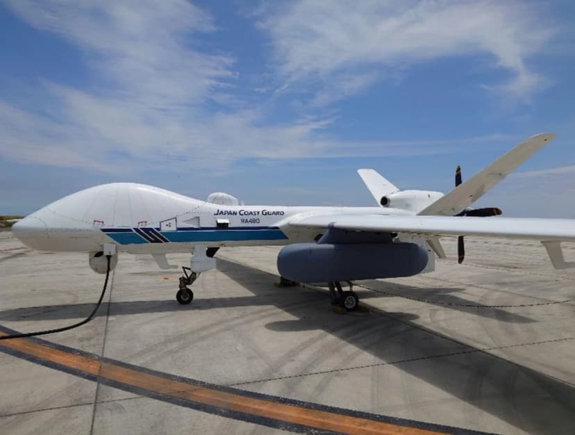滑走路に止まった白い大型の無人航空機。胴体に「JAPAN COAST GUARD」「RA480」の文字があり、胴体の下に灰色の細長い入れ物が取り付けられている