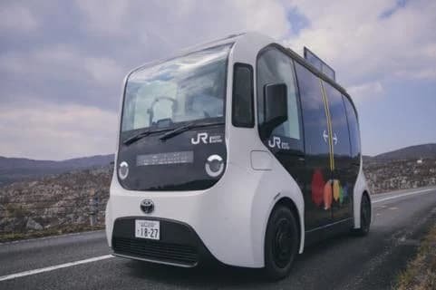 白い車体に黒いガラス面の、角の丸い小型の電気バス。前面と側面にJR西日本グループのマークがあり、車体の側面に色とりどりの丸い模様がある。海辺の道路を走る場面の写真